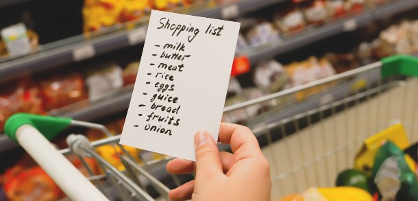 Hand holding a hand-written shopping list above a trolley with shelves of food in the background.