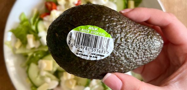 A hand holding an avocado with a date label on it with a bowl of salad in the background