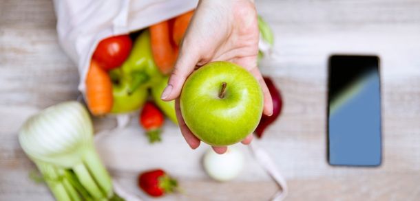 Hand holding a green apple above a bag of food next to a mobile phone.