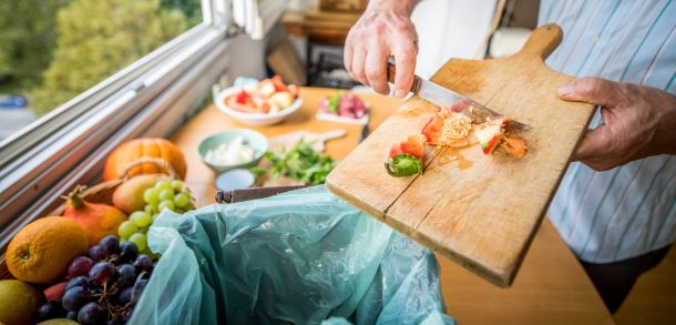 Hand scraping vegetable peelings into a food caddy with plates of food in the background
