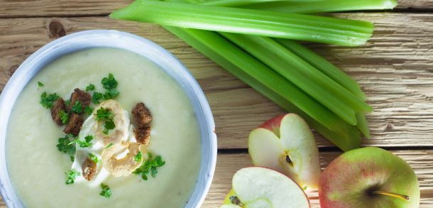 A bowl with apple soup, some apples and some celery on a wooden table. 