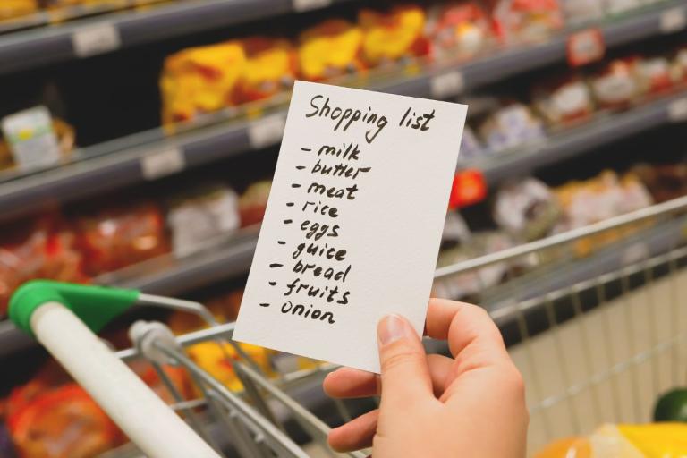 Hand holding a shopping list above a trolley in a shop.