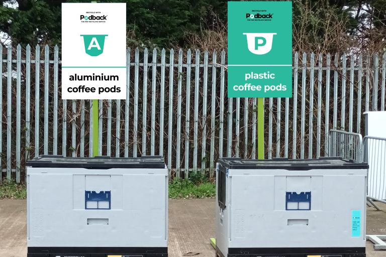 Two large, grey containers each with a rectangular sign above them. The white sign on the left says, “Recycle with Podback, The Pod Recycling Service: Aluminium Coffee Pods”. The turquoise sign on the right says, “Recycle with Podback, The Pod Recycling Service: Plastic Coffee Pods”. 