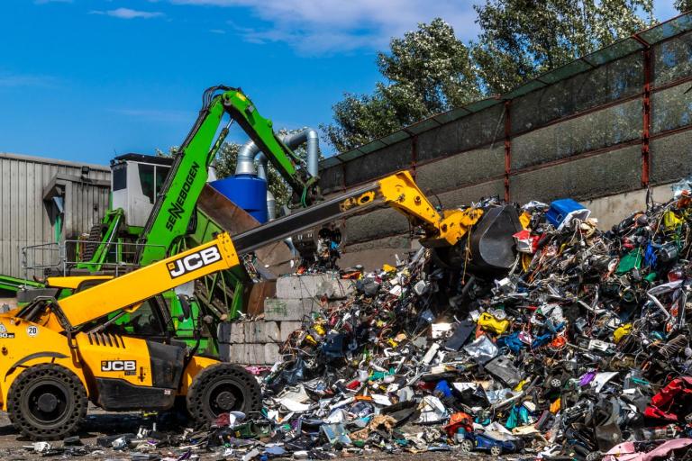 A yellow JCB sorting through a large pile of rubbish on the ground at a waste facility with a blue sky in the background.