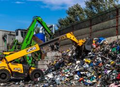 A yellow JCB sorting through a large pile of rubbish on the ground at a waste facility with a blue sky in the background.